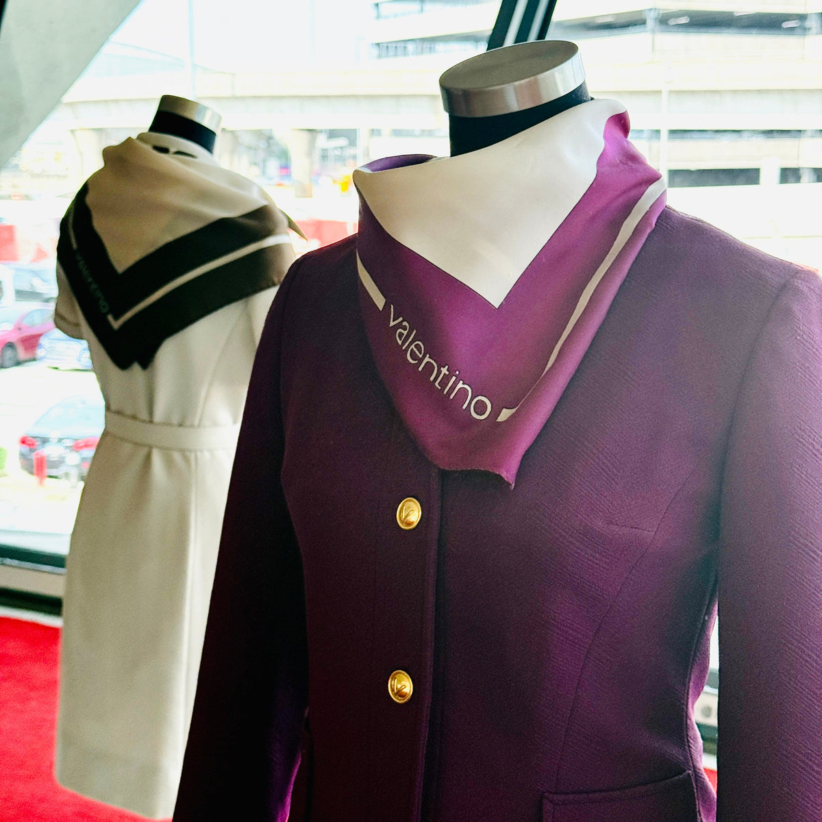 A purple Valentino TWA vintage uniform 
blazer with gold buttons displayed on a mannequin, paired with a matching purple neck scarf featuring the Valentino logo. A second cream-colored dress on a mannequin is visible in the background near a window.